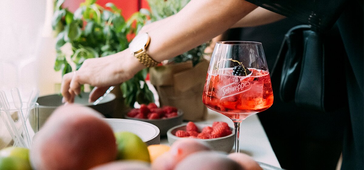 Eine Frau garniert einen Cocktail mit verschiedenen Früchten die Schälchen bereitstehen. 